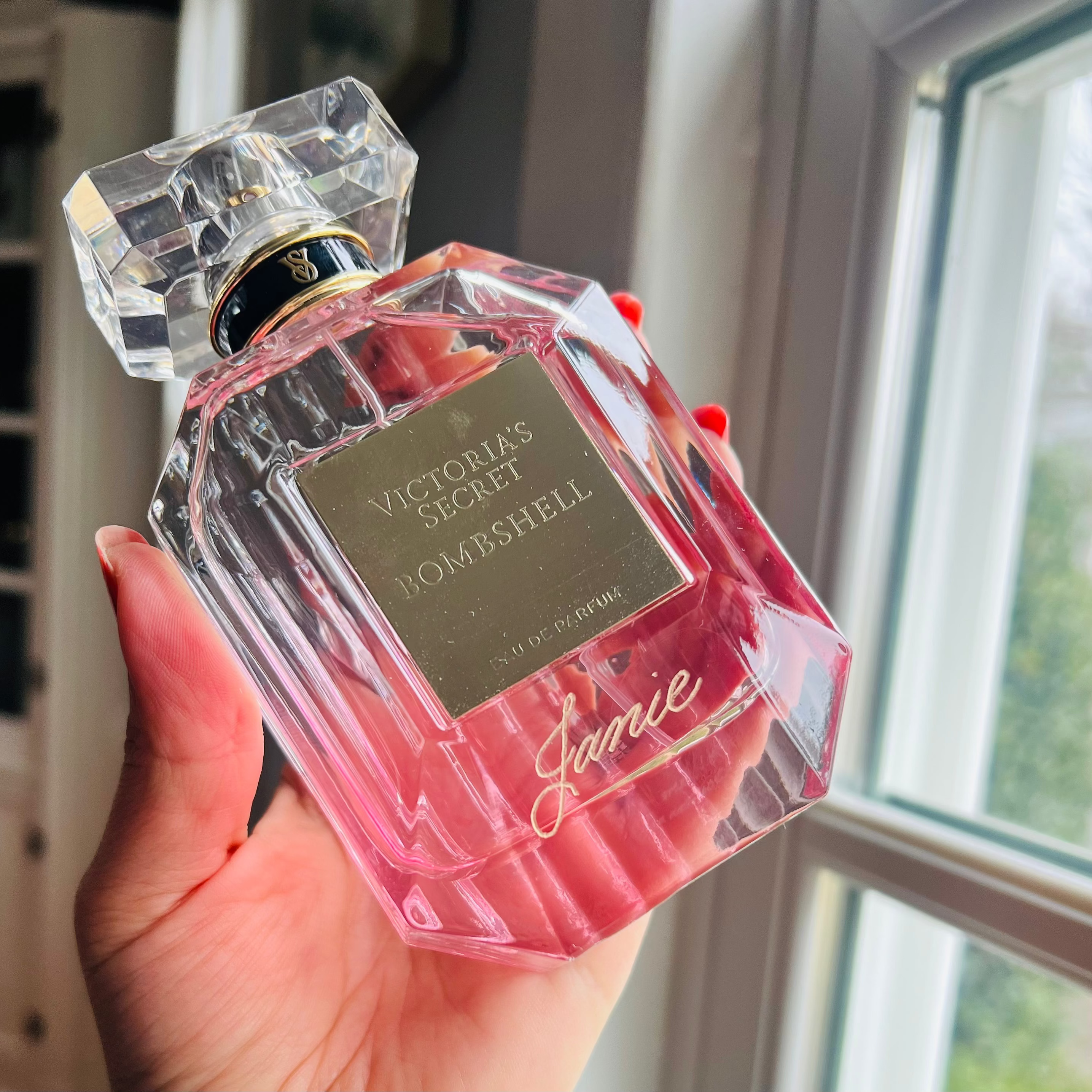 Pink perfume bottle with clear crystal design held by a hand, against a neutral indoor background.